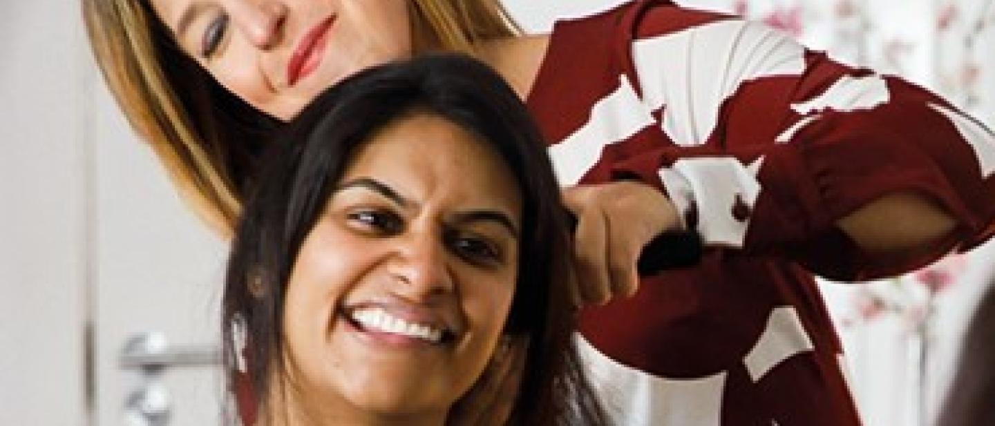 A woman smiles while having her hair styled by another woman in a brightly lit room, both reflected in a mirror.